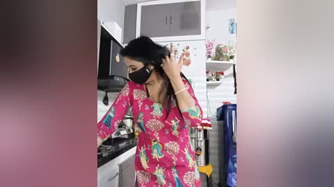 A video of a woman in a bright pink, patterned dress with a floral design, wearing a black face mask, fixing her hair in a kitchen with white cabinets, a stove, and colorful decor.