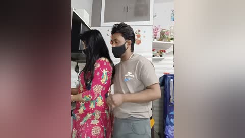 A video shows a woman in a colorful floral dress and a man in a beige shirt and gray pants, both wearing masks, in a modern kitchen with white cabinets and a blue recycling bin.
