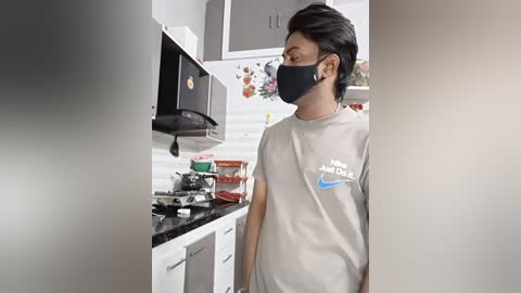 A video of a young man with short black hair and a black face mask, wearing a gray Nike t-shirt, standing in a modern kitchen with white cabinets and a black microwave.