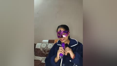 Video of a smiling woman with long black hair, wearing a purple mask, holding a purple vibrator, in a room with beige walls and a bed with floral-patterned sheets.