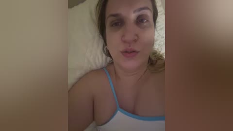 Video of a young woman with medium skin tone and brown hair, wearing a white tank top, lying on a bed with white sheets.