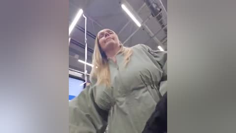 Video of a blonde woman with glasses, wearing a gray jacket, leaning forward in a dimly lit industrial space with metal beams and fluorescent lights.