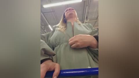 A low-angle video of a blonde woman in a light green coat, with a blue bar on the left, in an industrial setting with metal beams and fluorescent lights.