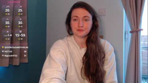 A video of a young woman with long brown hair, fair skin, and a neutral expression, wearing a white robe, standing indoors next to a window with beige curtains.