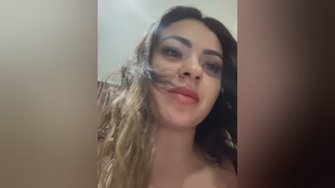 A video of a young woman with wavy, shoulder-length brown hair, fair skin, and a neutral expression. She is indoors, with the background blurred and out of focus.