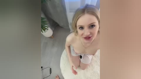 A video of a blonde woman in lingerie, kneeling on a white carpet, with a grey curtain and potted plant in the background.