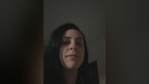 Video of a young woman with long black hair, light skin, and minimal makeup, smiling slightly. She is indoors with dim lighting, creating a moody atmosphere.