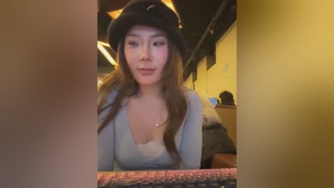 Video of an East Asian woman with long brown hair, wearing a black hat and light blue V-neck top, seated indoors with a colorful blanket.