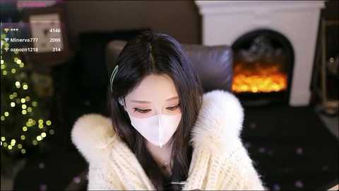 A video of an East Asian woman with long black hair, wearing a white face mask and a cream-colored, knitted sweater, sitting indoors near a lit fireplace.