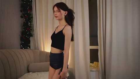 Media: Video of a slender, fair-skinned young woman with long brown hair, wearing a black lace bralette and high-waisted shorts, standing indoors by a beige couch, Christmas decorations in the background.