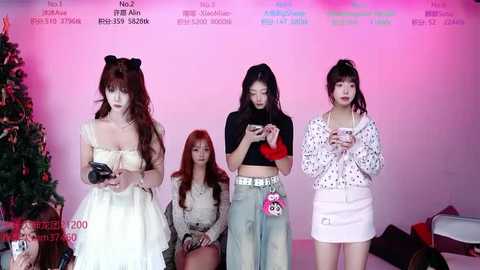 Video of four young women, two with long hair in white dresses, two in casual outfits, seated in a pink-lit room.