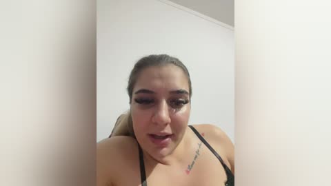 Video of a light-skinned woman with long brown hair, wearing black lingerie, standing in a dimly lit room with white walls and a noticeable shadow on her face.