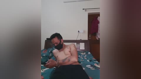 Video of a shirtless, light-skinned man with a small tattoo on his arm, wearing a face mask, lying on a bed with a colorful duvet, in a dimly lit, simple bedroom.