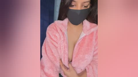 Video of a light-skinned woman with long dark hair, wearing a pink fluffy robe and black face mask, partially unzipped to reveal a white bra, in a dimly lit room with blue walls.