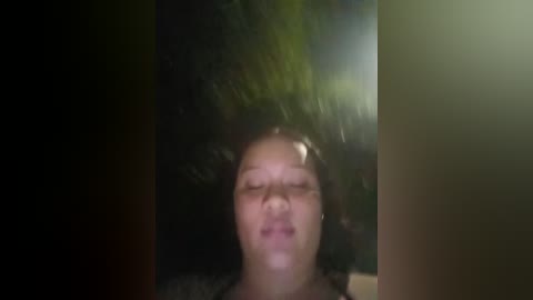 A video of a person with closed eyes, likely female, standing under a heavy rainfall, with blurred green foliage in the background.