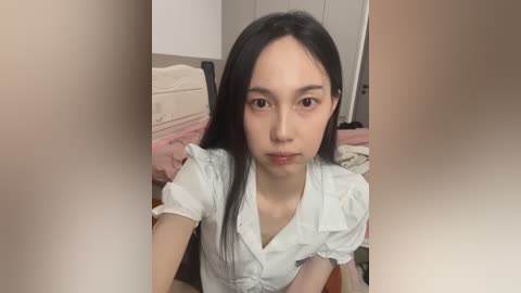 Video of a young Asian woman with straight black hair, wearing a white button-up shirt, seated in a bedroom with pink bedding and white walls.