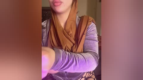 Video of a woman with light skin, wearing a gray long-sleeve top and a brown headscarf, arms crossed, lips slightly parted, in a dimly lit room.