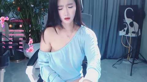Video of an East Asian woman with long black hair, wearing a light blue off-shoulder top, seated in a modern room with greenery, pink lighting, and abstract art on the wall.
