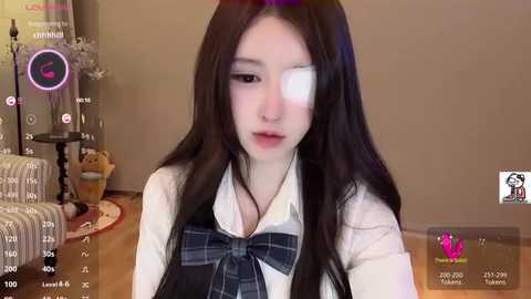 A video of an Asian woman with long black hair, wearing a white shirt and plaid vest, with a black eye patch, sitting in a cozy, beige-toned living room.