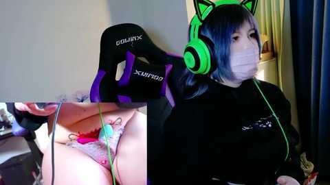 Video collage featuring a young woman with light skin and blue hair, wearing green cat ear headphones, a black top, and a face mask, sitting at a gaming chair in a dimly lit room.