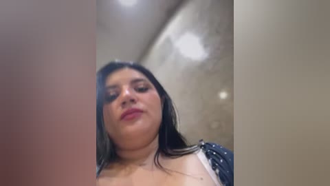 Video of a plus-size Latina woman with long black hair, wearing a white top, and a navy blue jacket, standing in a dimly lit, beige-toned hallway.