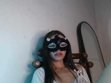 Video of a woman with long black hair, wearing a black lace mask and a white top, seated against a plain white wall.