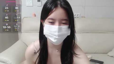 Video of an Asian woman with long black hair, wearing a white mask and a light-colored top, sitting on a white couch, in a sparsely decorated room.