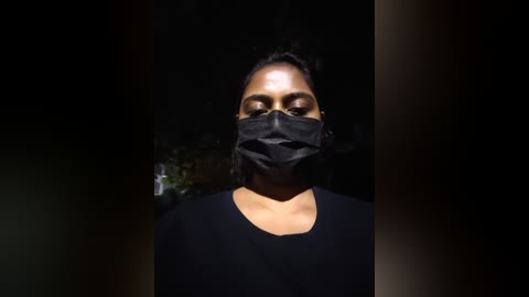 Video of a person with medium-brown skin, wearing a black mask and shirt, standing in a dark, dimly lit outdoor setting with indistinct background foliage.