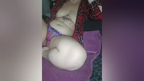 A video of a plus-size woman with fair skin, lying on a purple towel, wearing a red plaid shirt and blue floral panties. Her tattooed abdomen is visible.