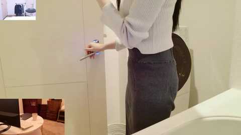 Media: Video of a woman in a light blue sweater and high-waisted gray skirt, holding a metal object, standing in a modern, minimalist bathroom with a white sink and mirror.