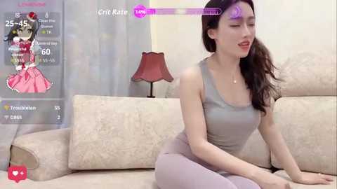 A video of a young Asian woman with long dark hair, wearing a grey tank top and leggings, sitting on a beige couch in a modern living room.