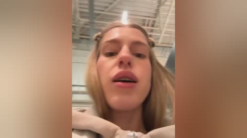 A close-up video of a blonde woman with straight hair, light skin, and slightly parted lips. The background shows a white ceiling and walls in a modern room.