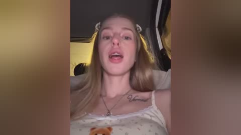 Video of a young, fair-skinned woman with blonde pigtails, wearing a white spaghetti-strap top and a necklace. She's sitting in a dimly lit room with a yellowish hue, possibly a car.