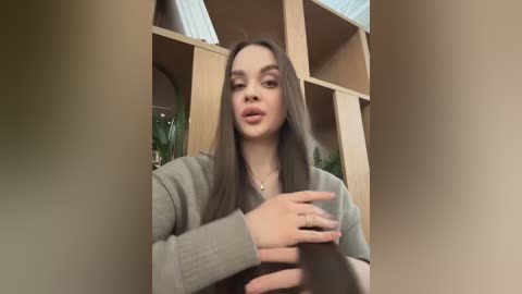 Video of a young woman with long brown hair, fair skin, and a neutral expression, wearing a beige cardigan, standing in front of a wooden bookshelf with green plants in the background.