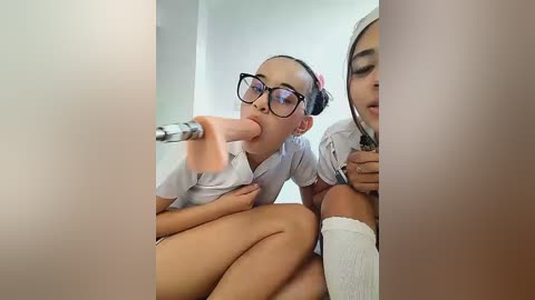 Video of two young women in school uniforms, one performing oral sex on a large, erect penis. The background is minimalistic and white, focusing attention on the explicit act.