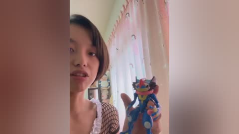 Video of a young Asian woman with short dark hair, wearing a patterned top, standing in a room with a pink curtain and a colorful dragon toy.