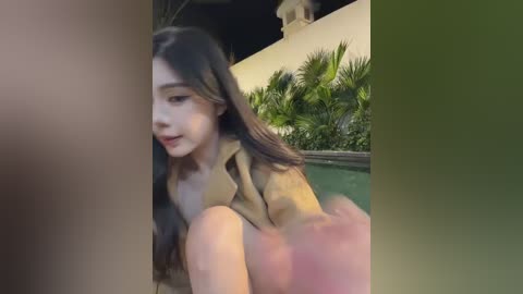 A blurred video of a young woman with long, dark hair, wearing a beige jacket, sitting by a poolside at night. The background features lush green plants and a beige wall.