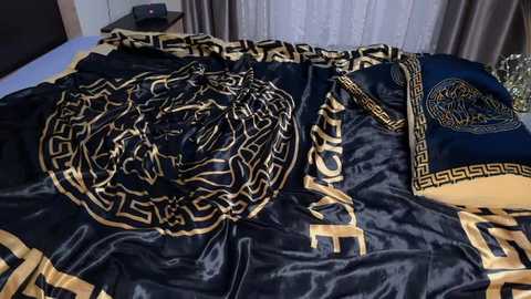 Video of a bed covered in luxurious, dark blue satin sheets with intricate gold Greek key patterns and a matching pillowcase. The bedding is neatly arranged, with a hint of a bedside table visible in the background.