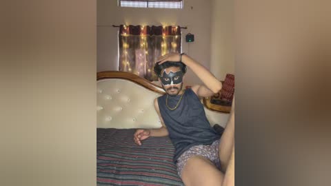 A video of a man in a dark room, wearing a black mask, sleeveless shirt, and patterned shorts, sitting on a bed with striped sheets, looking at the camera.