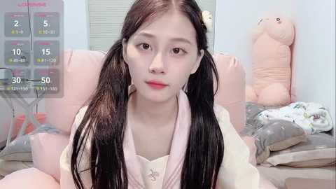 Video of a young East Asian woman with long black hair in pigtails, wearing a light pink robe, sitting on a bed with plush toys, in a pastel-colored room with a digital clock displaying 30 minutes.