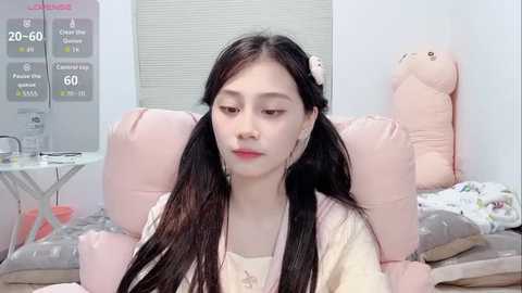 Video of a young Asian woman with long black hair in pigtails, wearing a beige robe, sitting on a bed with plush pink pillows and a stuffed animal in a pastel room.