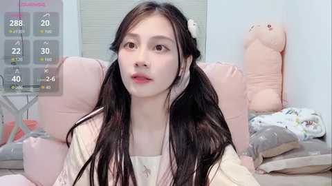 A video of an East Asian woman with long black pigtails, fair skin, and light makeup, sitting on a pastel pink chair in a cozy, girly room with plush toys and a digital clock.
