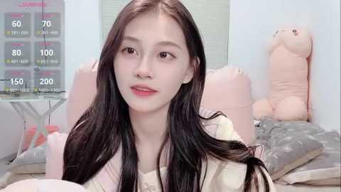 Video of an East Asian woman with long black hair, fair skin, wearing a white top, sitting on a bed with pastel-colored pillows and a plush toy.