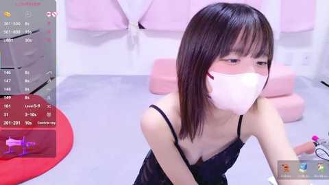 Video of an East Asian woman with short black hair, wearing a pink face mask and black lace lingerie, kneeling in a pink room.