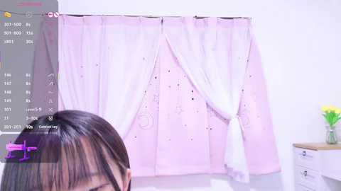 A video of a young Asian woman with straight black hair, wearing a white top and pink headband, stands in front of a pink curtain with white polka dots. The room is bright with a white wall and a wooden nightstand with yellow flowers in a vase.