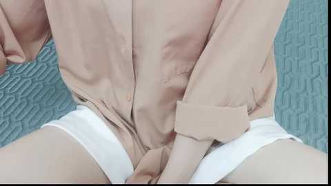 Video of a person's torso in beige clothing with a zipper down the middle, revealing white underwear. Background shows a teal, quilted bedspread.