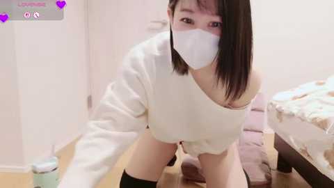 Video of an East Asian woman with short black hair and a white mask, wearing a white off-shoulder sweater, black stockings, and no pants, leaning over a table in a dimly lit bedroom with a bed and wooden floor.