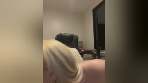 Video of a blonde woman, viewed from behind, sitting on a black leather office chair in a dimly lit room with beige walls.