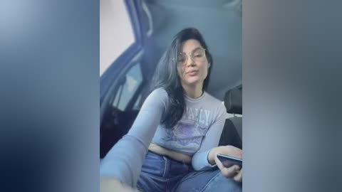 A candid video of a young woman with long black hair, wearing a light grey graphic t-shirt and blue jeans, sitting in the back seat of a car, looking at her smartphone.