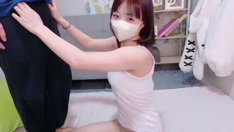 Video of an Asian woman with straight brown hair and a white mask kneeling, wearing a white tank top and pants, holding a man's leg in a bathroom with white towels and a green towel on the floor.
